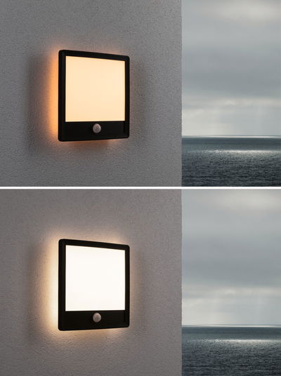 Black square outdoor wall light with motion sensor and warm white illumination on grey wall.