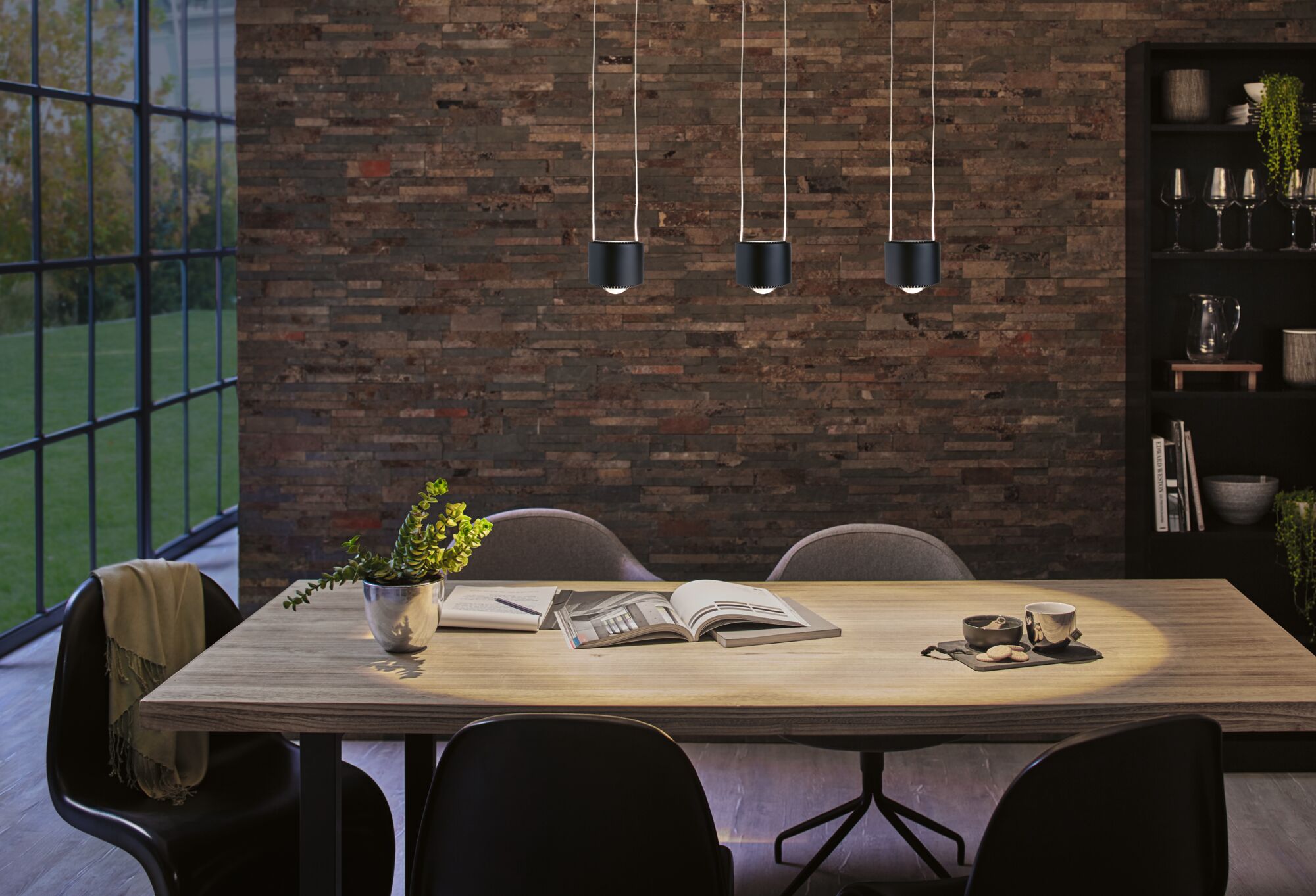 Three black metal pendant lights above light wooden table with books and cup in modern dining room.