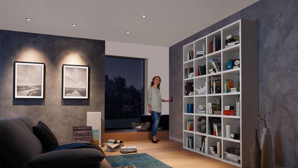 Modernes Wohnzimmer mit blendfreier LED-Deckenbeleuchtung, grauen Wänden und weißem Bücherregal mit Büchern.