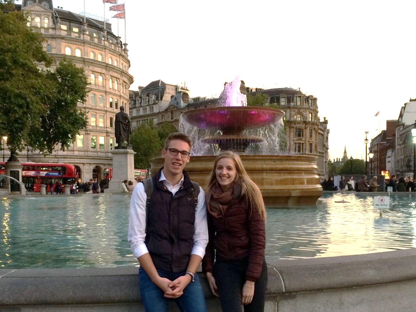 Zwei junge Menschen vor beleuchtetem Springbrunnen in London mit roten Doppeldeckerbussen im Hintergrund.