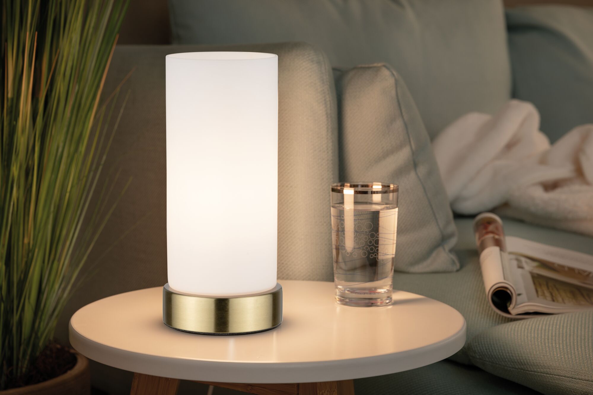 Moderne Tischlampe mit weißem Glas und Messingfuß auf rundem Beistelltisch neben Glas Wasser