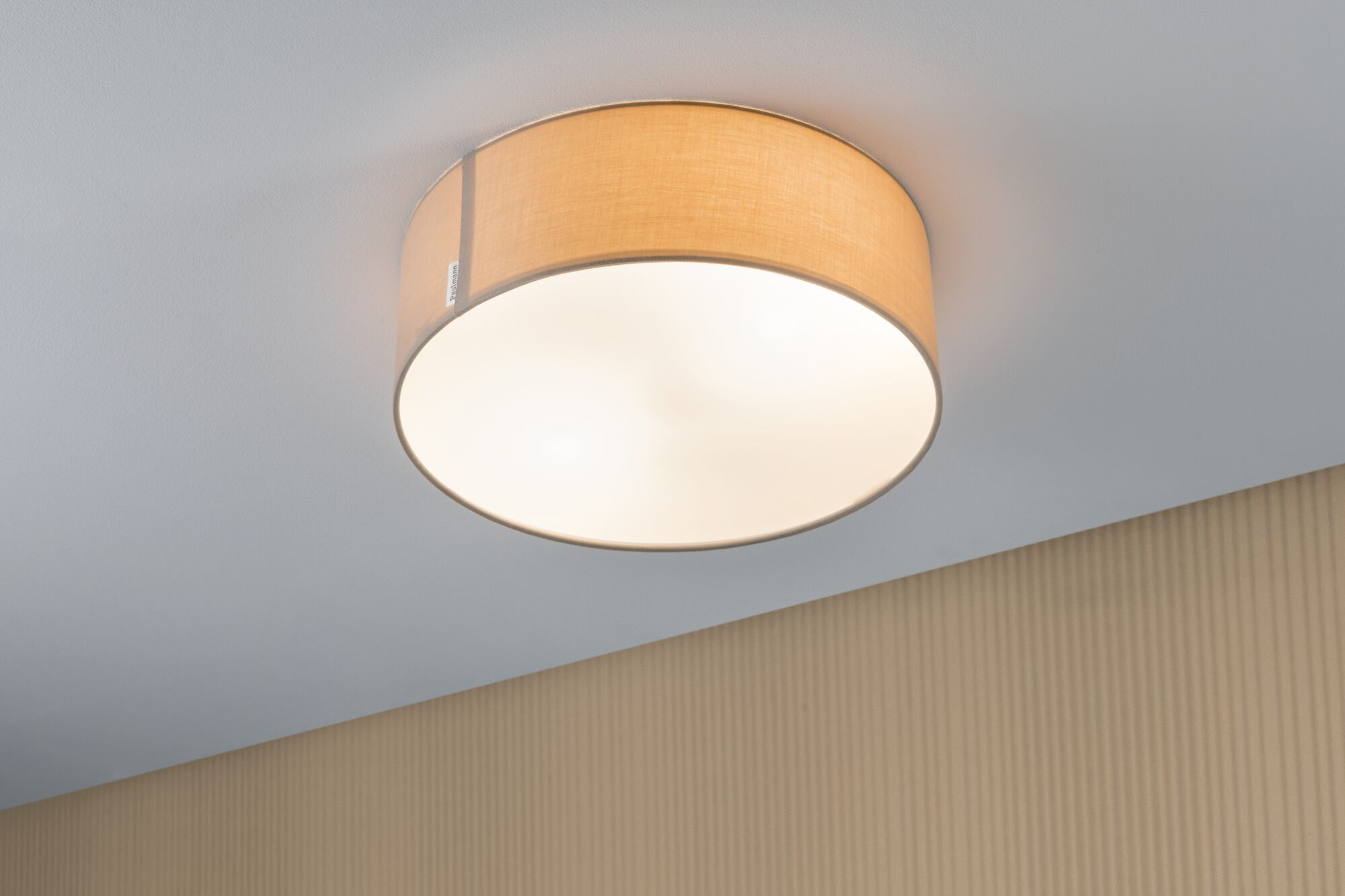 Rund loftlampe med beige stofskærm og hvid diffusor, der giver varmt lys