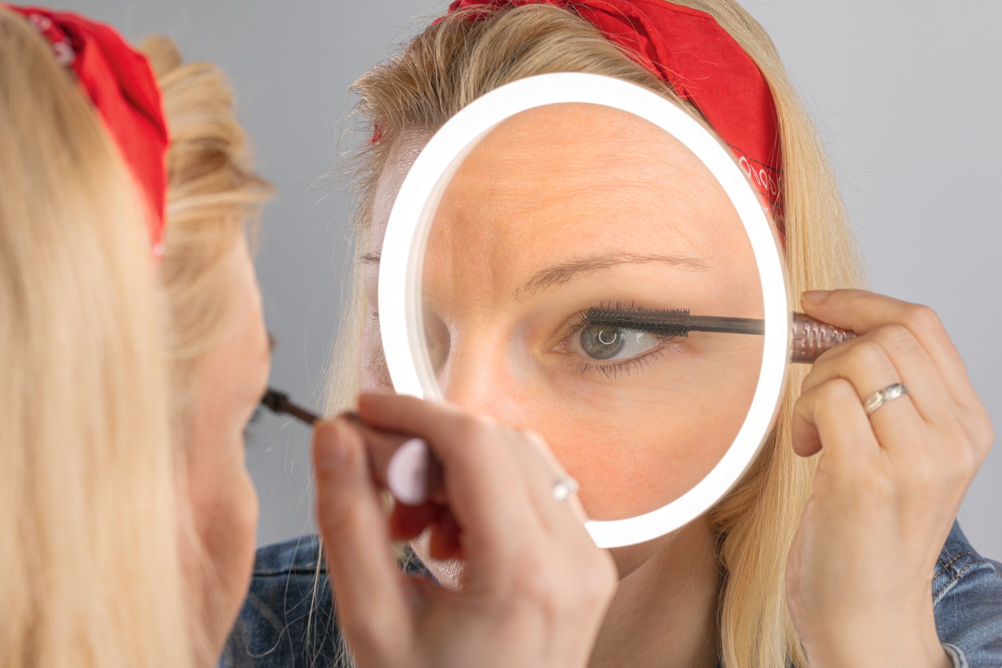 Woman applying black mascara using illuminated LED ring cosmetic mirror for precise makeup.