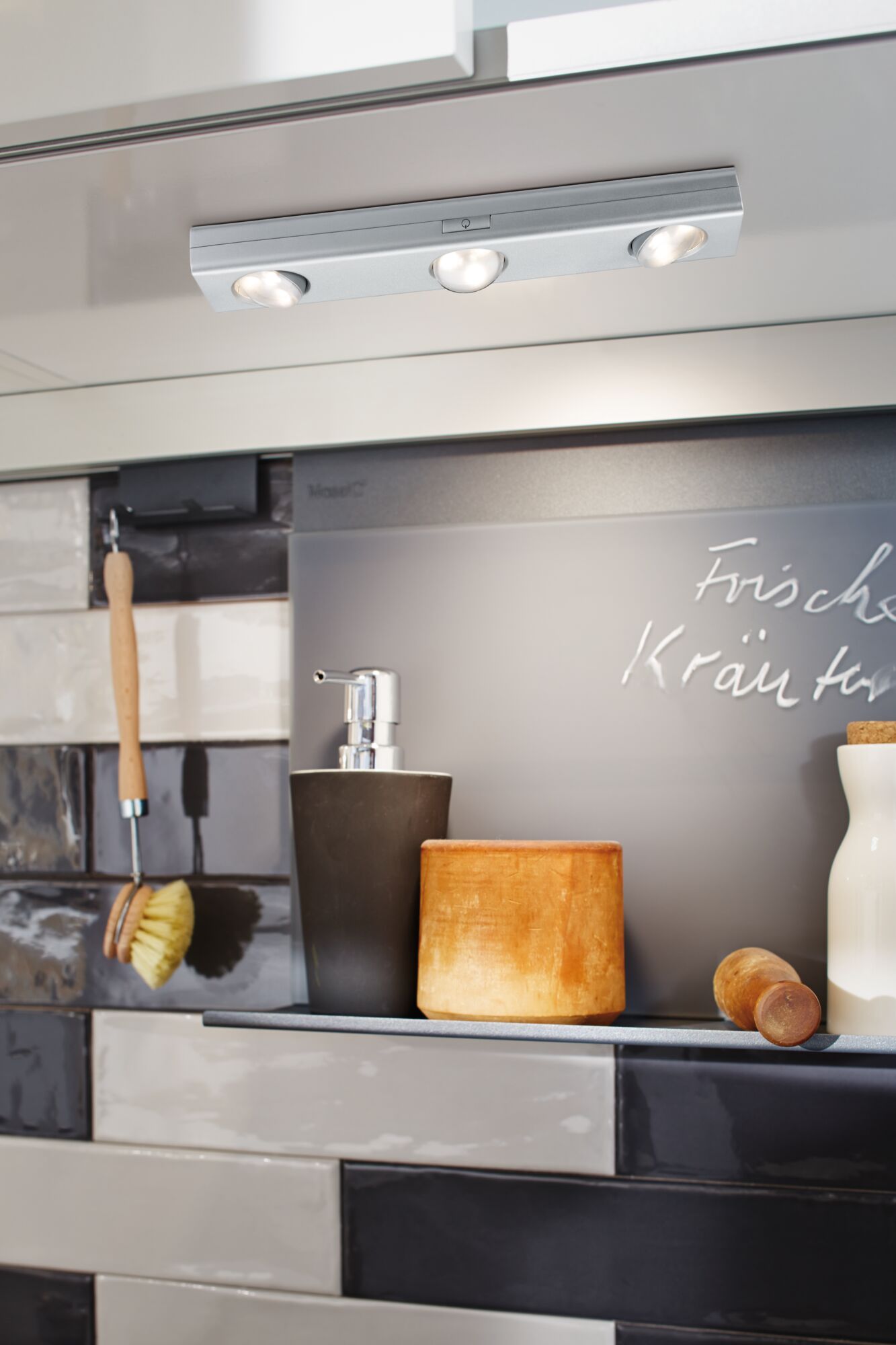 Trois lampes LED métalliques argentées sous meuble au-dessus d'étagère de cuisine avec distributeur de savon et objets en bois.