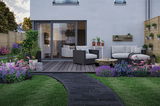 Jardin moderne avec terrasse en bois grise, mobilier lounge noir et parterres de fleurs colorées pour éclairage extérieur.