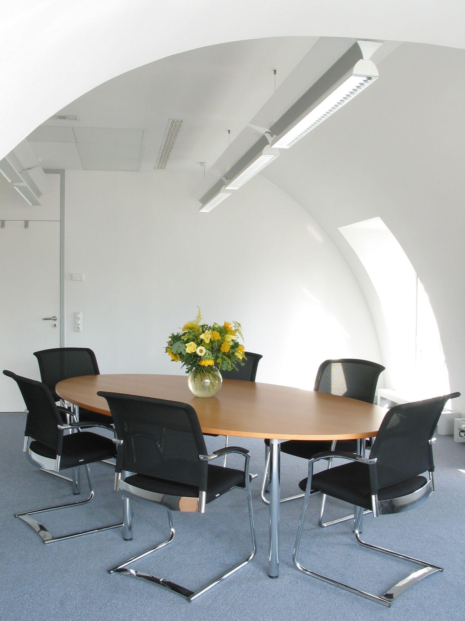 Moderner Konferenztisch mit Holzplatte und sechs schwarzen Mesh-Stühlen in hellem Büro mit LED-Deckenleuchten
