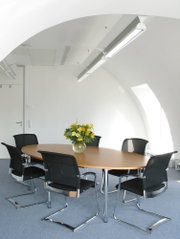 Moderne vergadertafel met houten blad en zes zwarte mesh stoelen in lichte kantoorruimte met LED-plafondlampen