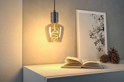 Hanging LED bulb with warm white light and grey metal holder above white sideboard with book and decor.