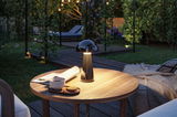 Schwarze LED-Tischleuchte aus Metall auf Holztisch im Garten mit Tasse, Keksen und Zeitung bei Abendlicht