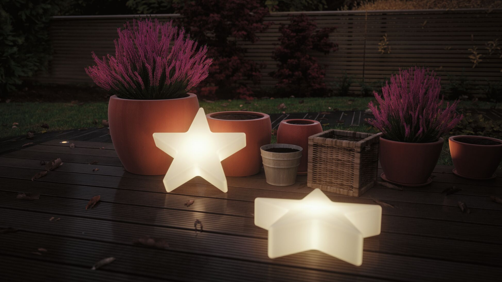 Zwei leuchtende Sternlampen auf Holzterrasse neben Terrakotta- und Korbblumentöpfen im Garten