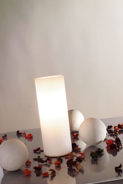 Cilindervormige witte glazen tafellamp met warm licht, omringd door decoratieve witte bollen en gedroogde bloemblaadjes