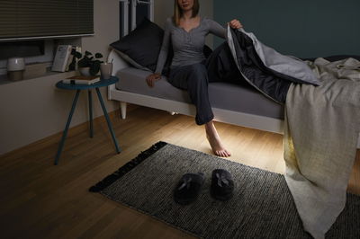 Femme assise sur lit gris avec couette noire, sol en bois éclairé avec chaussons gris et tapis
