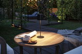 Lampe de table LED noire en métal sur table ronde en bois dans un jardin avec tasse et biscuits.