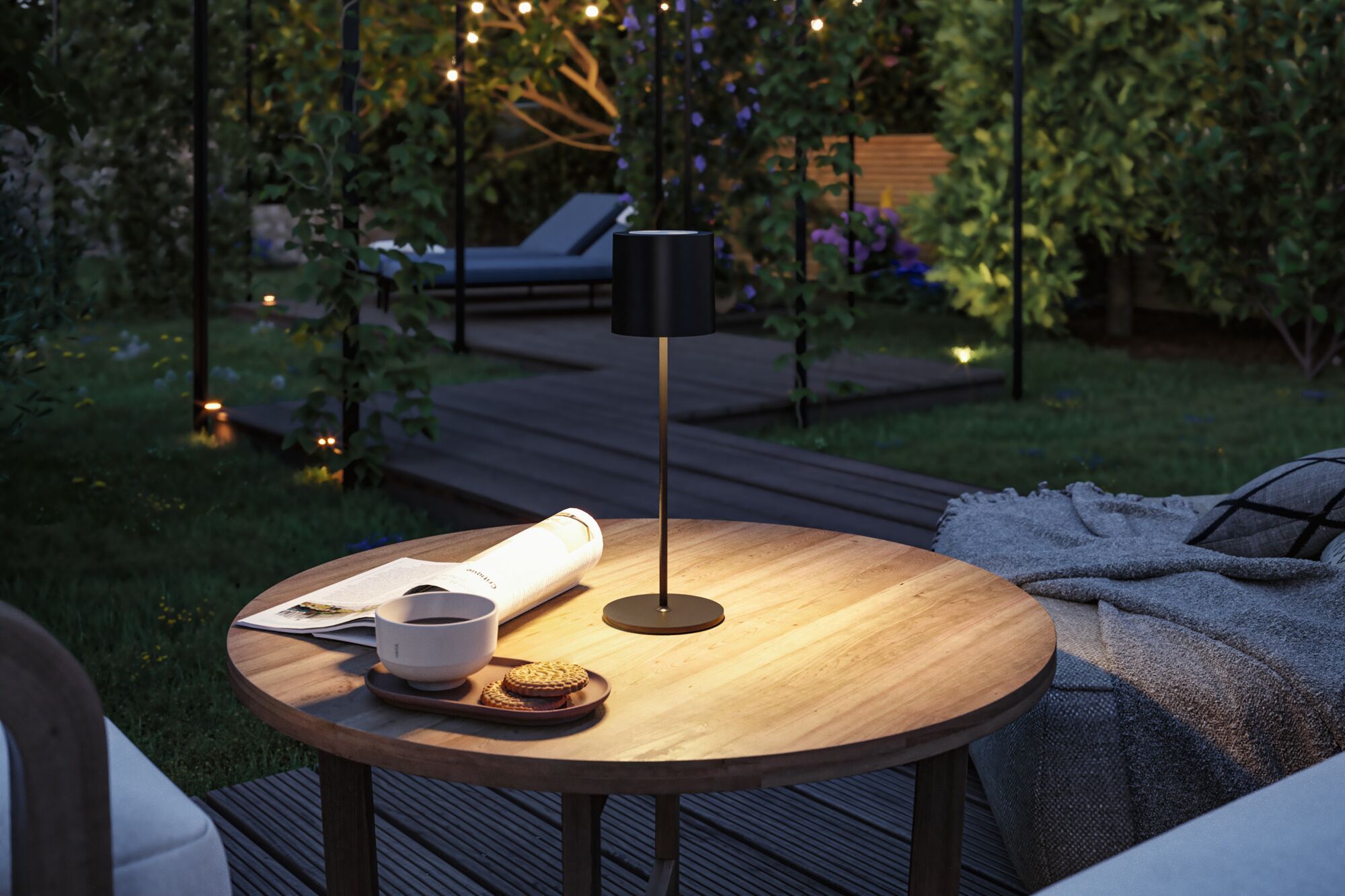 Black metal LED table lamp on round wooden garden table with cup and cookies in evening.