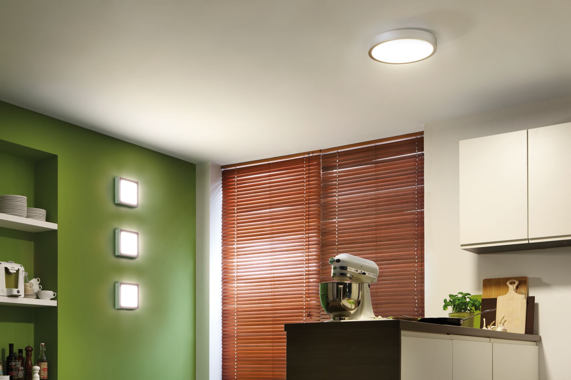 Modern round ceiling light and three square wall lamps in white in a green kitchen with wooden blinds.