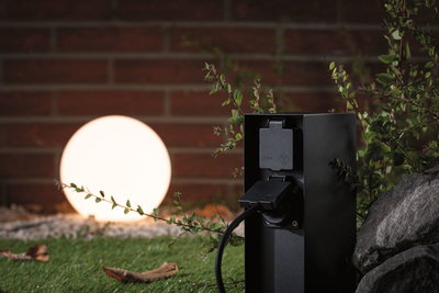 Zwarte buitenstekker met IP44 bescherming bij verlichte bolvormige tuinlamp op gras met stenen