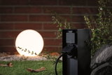 Prise extérieure noire IP44 devant une lampe sphérique lumineuse dans un jardin avec herbe et pierres