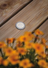 Round LED recessed floor light in brushed grey aluminum on wooden terrace with flowers.