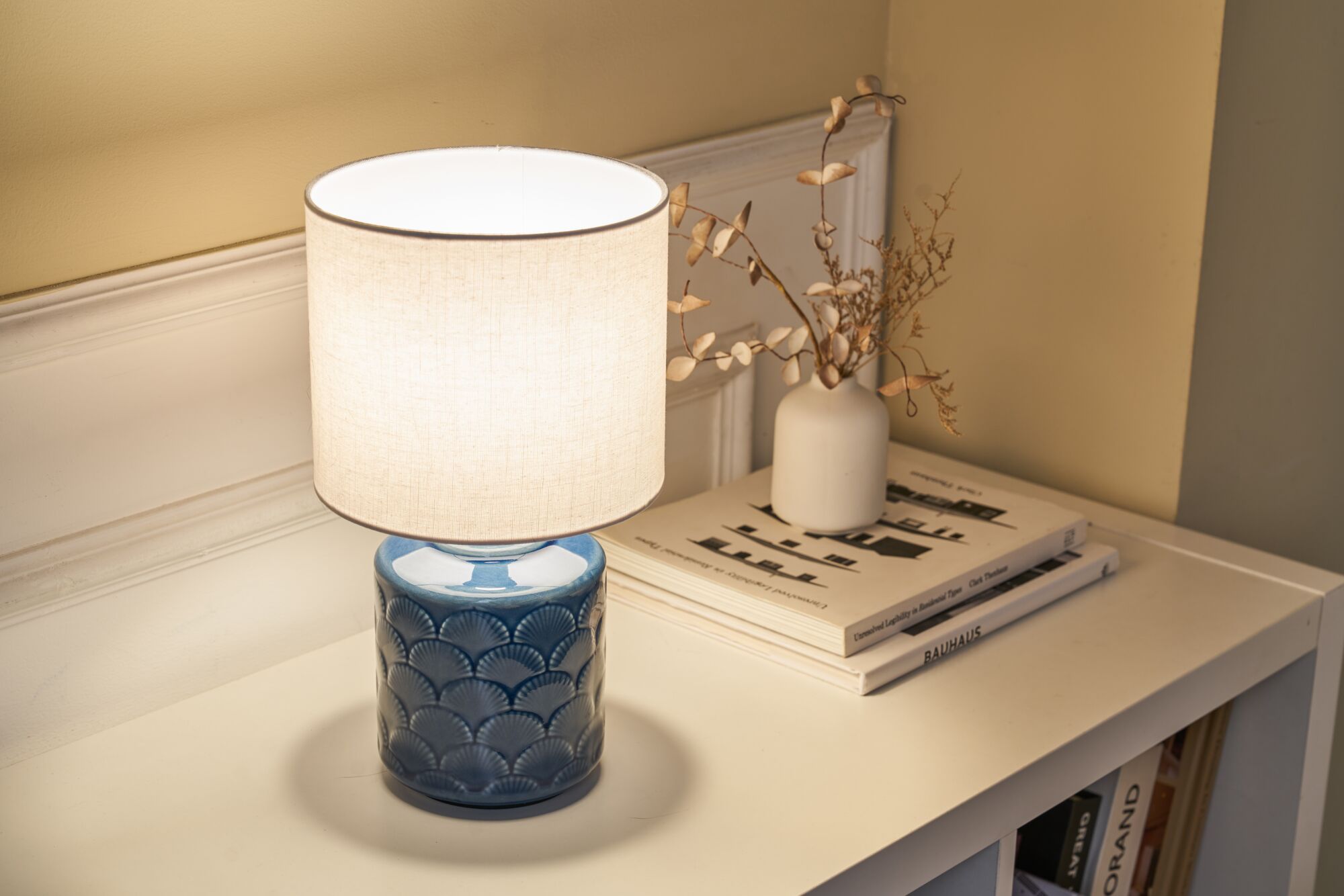 Tafellamp met blauwe keramische voet en witte stoffen kap op witte plank met decoratie en boeken