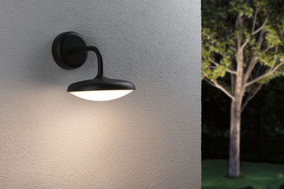 Schwarze Außenwandleuchte aus Metall mit warmweißem Licht an grauer Wand im Gartenbereich.