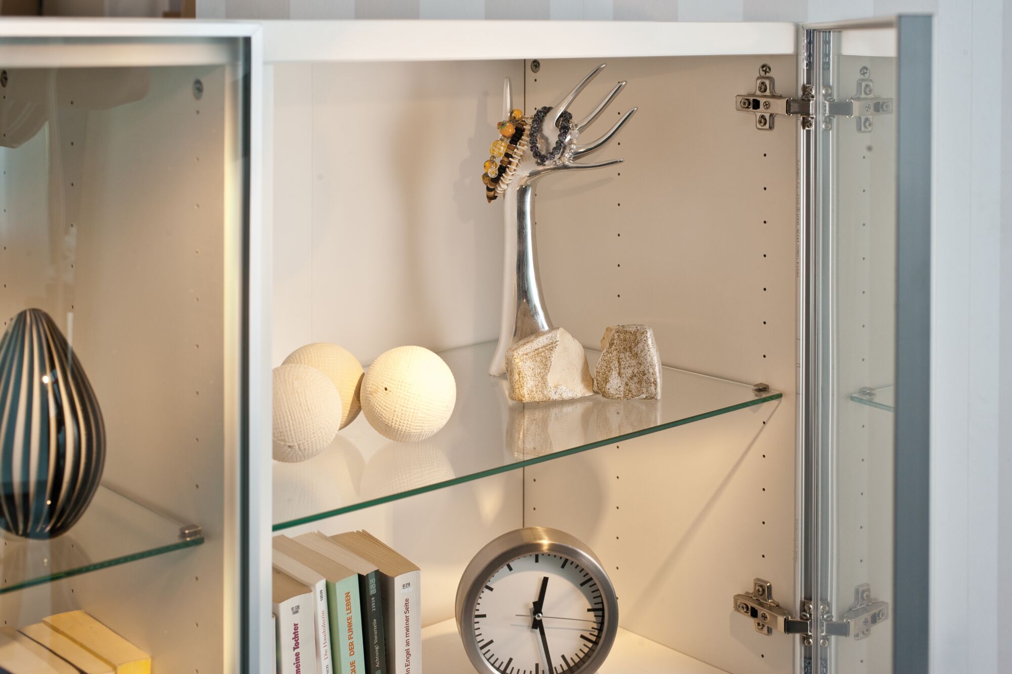 Weiße Dekokugeln und silberner Schmuckhalter in beleuchtetem Glasvitrinenschrank mit Uhr und Büchern