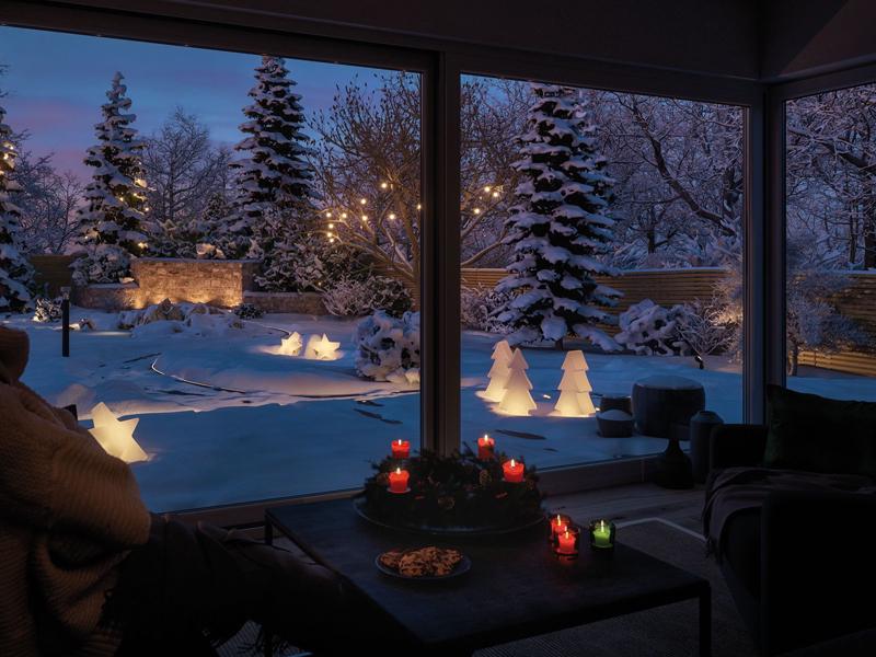 Wintergarten mit LED-Dekoleuchten in Form von Tannenbäumen und Sternen im verschneiten Garten bei Abenddämmerung
