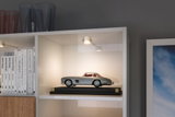 Small grey model car on black base lit by white LED ceiling spotlight in shelf display.