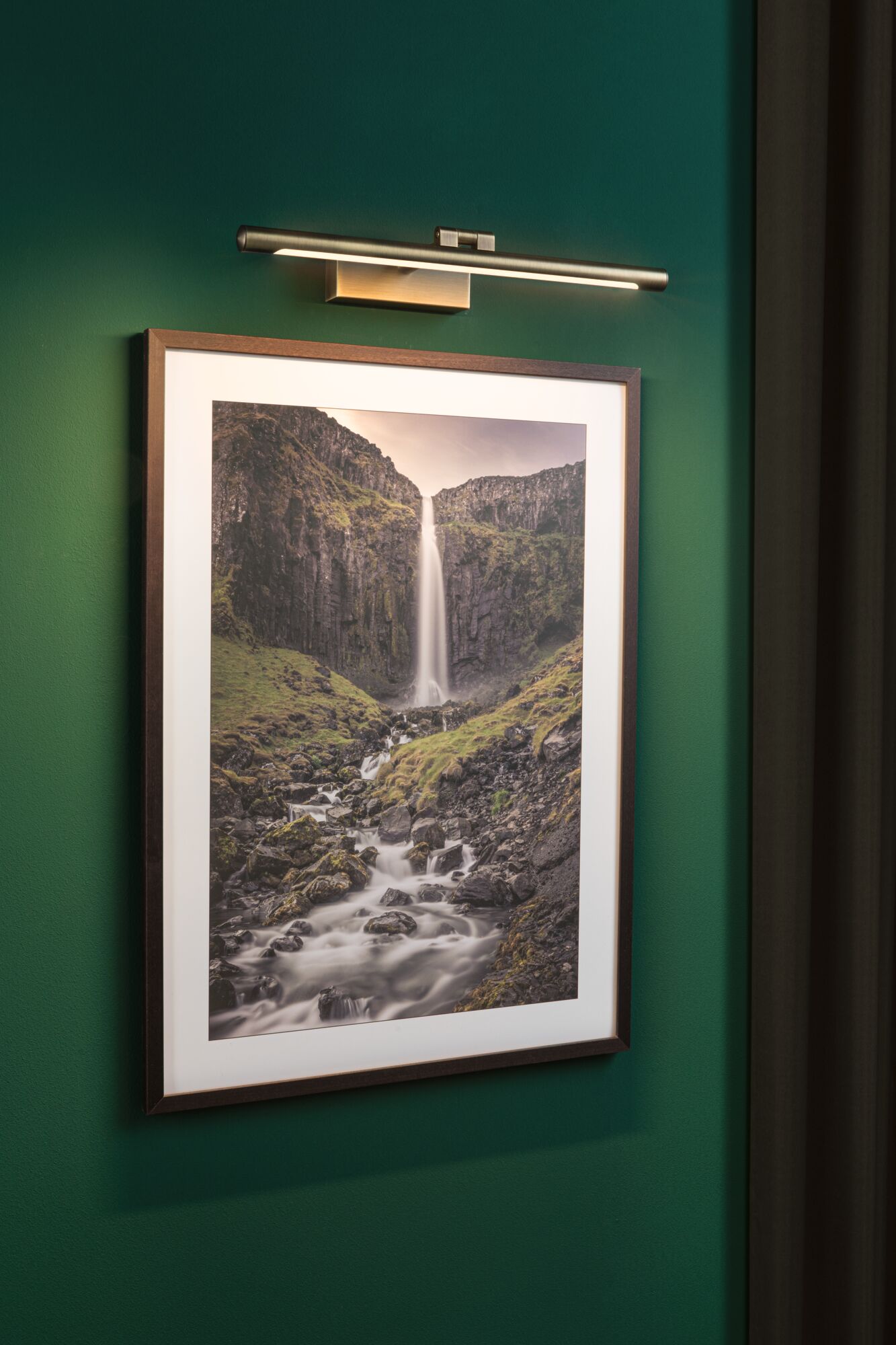Modern LED picture light in brushed metal on green wall above landscape photo with waterfall.