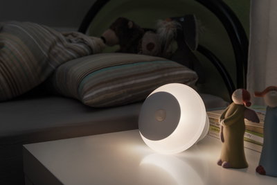 Round white LED night light with motion sensor on bedside table near children's bed and toys