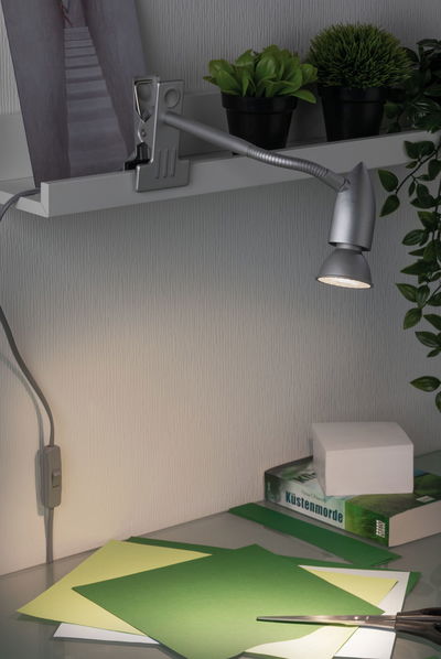 Silver metal LED clip lamp with flexible arm lighting green craft paper on desk for home office.