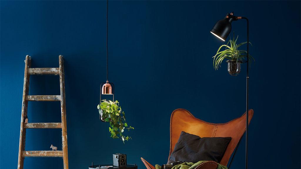 Wohnzimmer mit braunem Ledersessel, schwarzer Stehlampe mit Pflanze und hölzerner Leiter vor dunkelblauer Wand