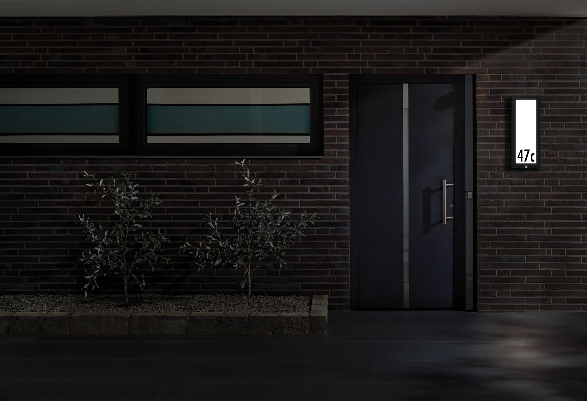 Modernes Haus mit dunkler Tür und beleuchtetem rechteckigem LED-Hausnummernschild an Backsteinwand