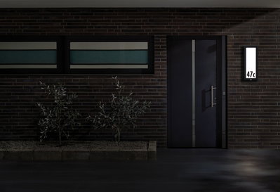 Modernes Haus mit dunkler Tür und beleuchtetem rechteckigem LED-Hausnummernschild an Backsteinwand