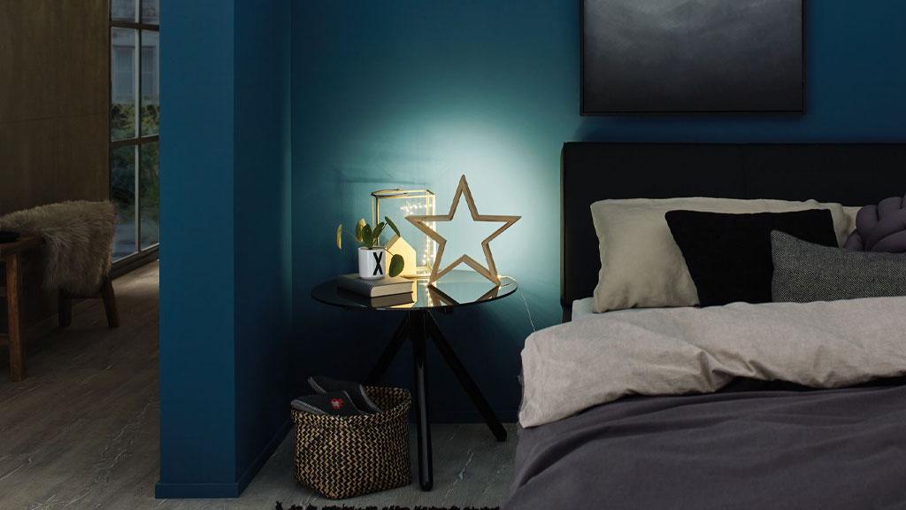 Schlafzimmer mit blauem Wandhintergrund, schwarzem Nachttisch und dekorativer Sternlampe aus Holz mit LED-Beleuchtung.