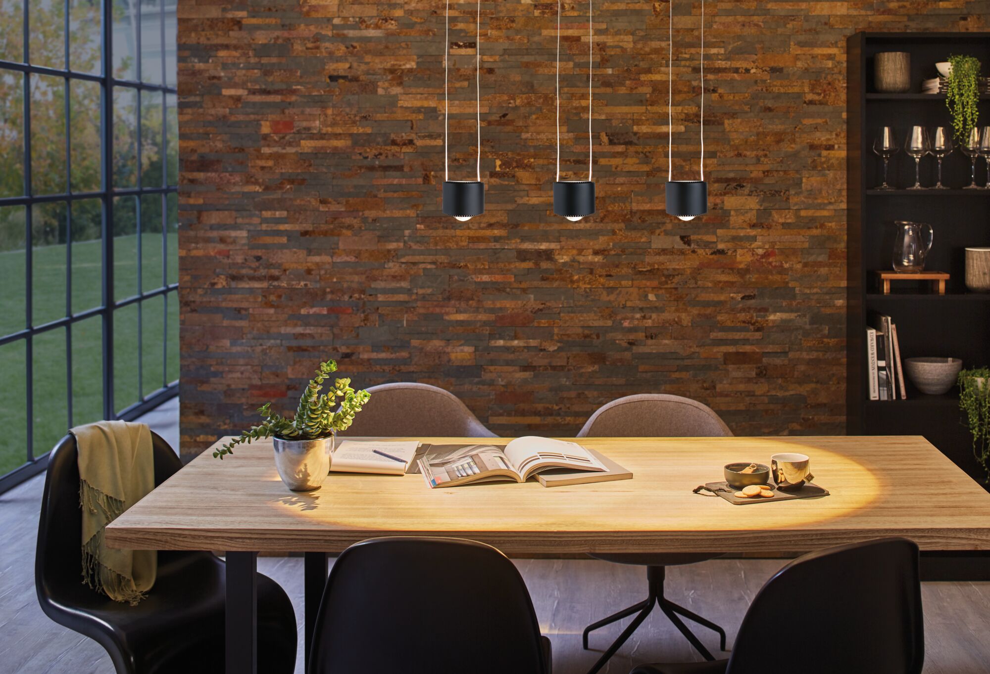 Modern dining area with wooden table, black chairs, and three black LED pendant lights on brick wall.