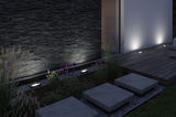 Modern outdoor floor lights illuminating dark stone wall and wooden terrace in garden at night.