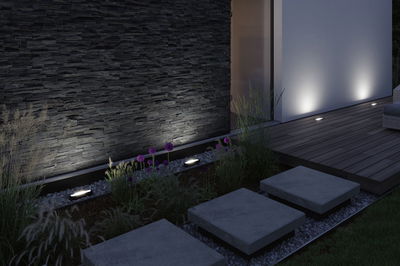 Moderne buitenverlichting met inbouwspots in de grond die donkere stenen muur en houten terras verlichten.