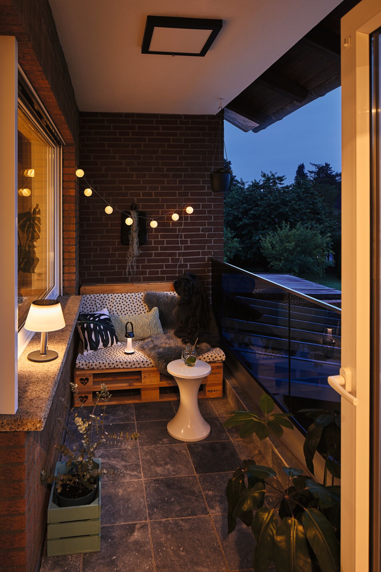 Cozy balcony with wooden pallet bench, white cushions, and warm white LED string lights for ambient lighting.