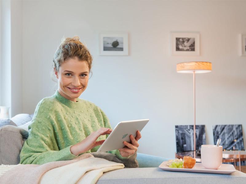 Frau mit grünem Pullover sitzt auf Sofa und nutzt Tablet, gemütliche Wohnzimmerszene mit Stehlampe.