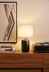 Modern table lamp with black metal base and white fabric shade on wooden sideboard next to turntable.