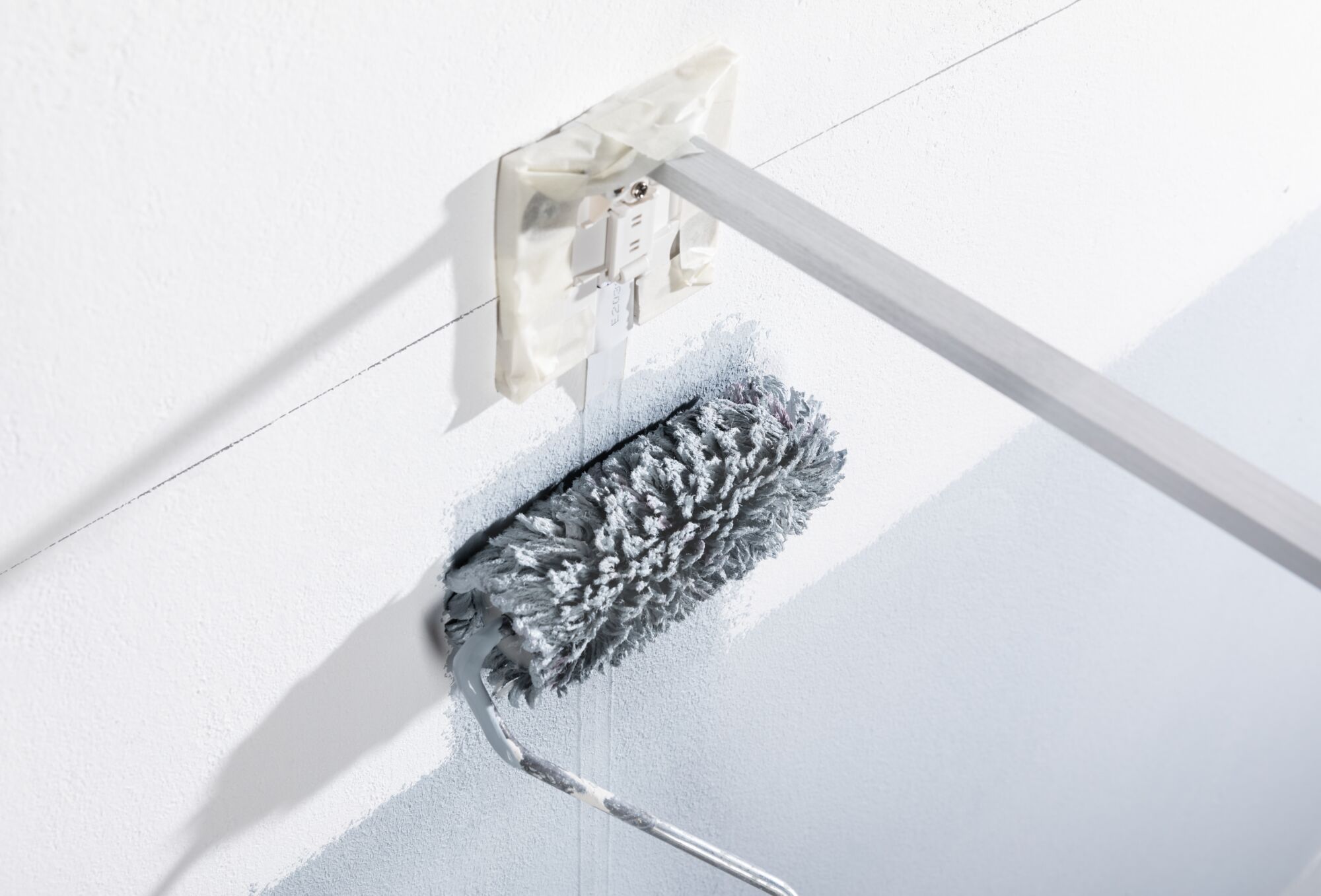Rouleau à peinture gris avec manche long peignant un mur blanc près d'une prise électrique