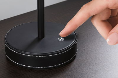 Base ronde noire de lampe en cuir avec couture blanche, doigt touchant un petit interrupteur sur surface en bois