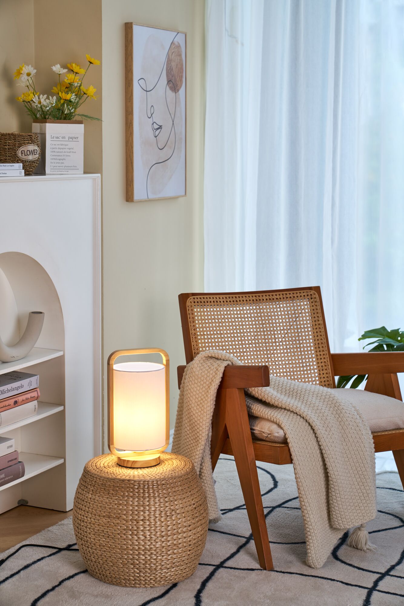 Moderne houten tafellamp met stoffen kap op gevlochten kruk naast houten stoel met deken in woonkamer