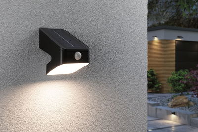 Black solar wall light with motion sensor on grey wall, warm white LED illumination for outdoor use.