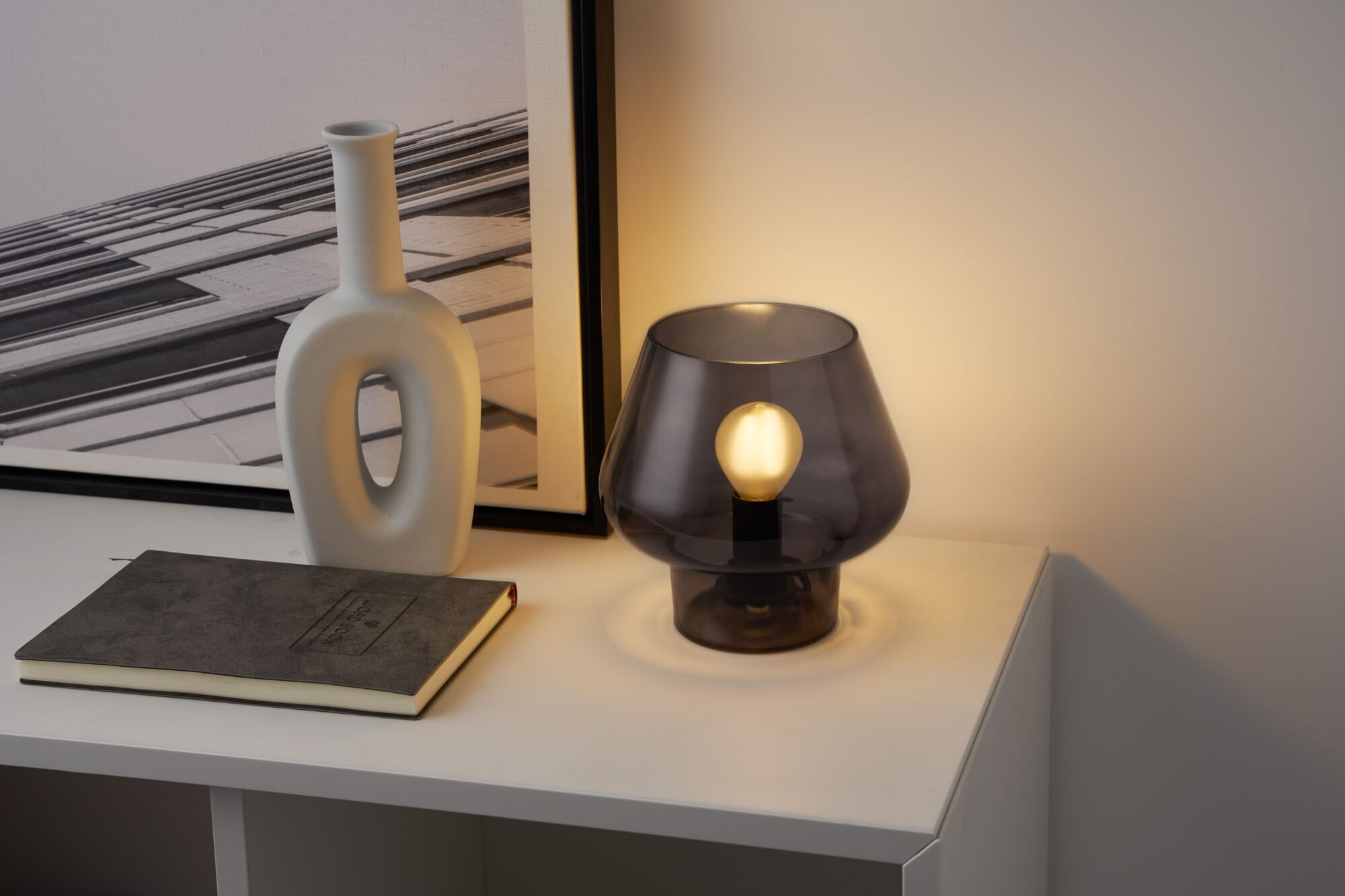 Moderne Tischleuchte aus rauchgrauem Glas mit sichtbarer Glühbirne auf weißem Sideboard neben Vase und Buch
