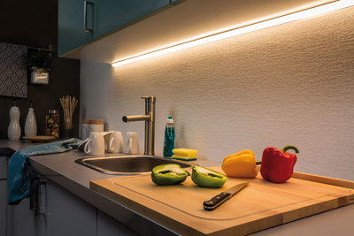 Plan de travail de cuisine moderne avec évier en acier inoxydable, planche à découper en bois et poivrons colorés sous lumière LED sous meuble
