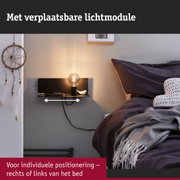 Zwarte wandlamp met verplaatsbare lichtmodule naast bed met donkere kussens en deken