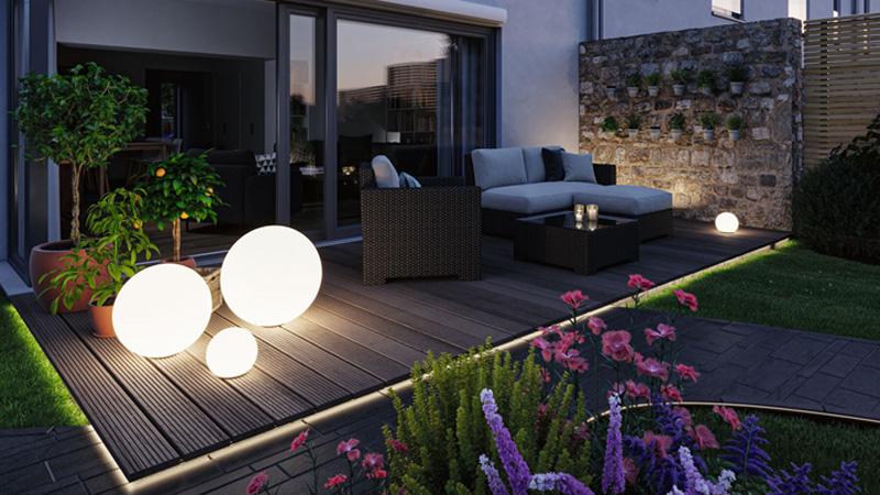 Moderne runde LED-Gartenleuchten in Weiß auf Holzterrasse mit Lounge-Möbeln und Blumenbeet bei Abendlicht
