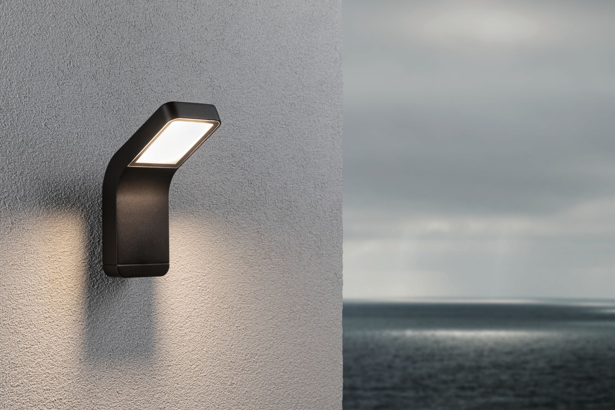 Schwarze LED-Außenwandleuchte aus Metall mit warmweißem Licht an grauer Wand mit Meerblick