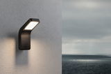 Schwarze LED-Außenwandleuchte aus Metall mit warmweißem Licht an grauer Wand mit Meerblick